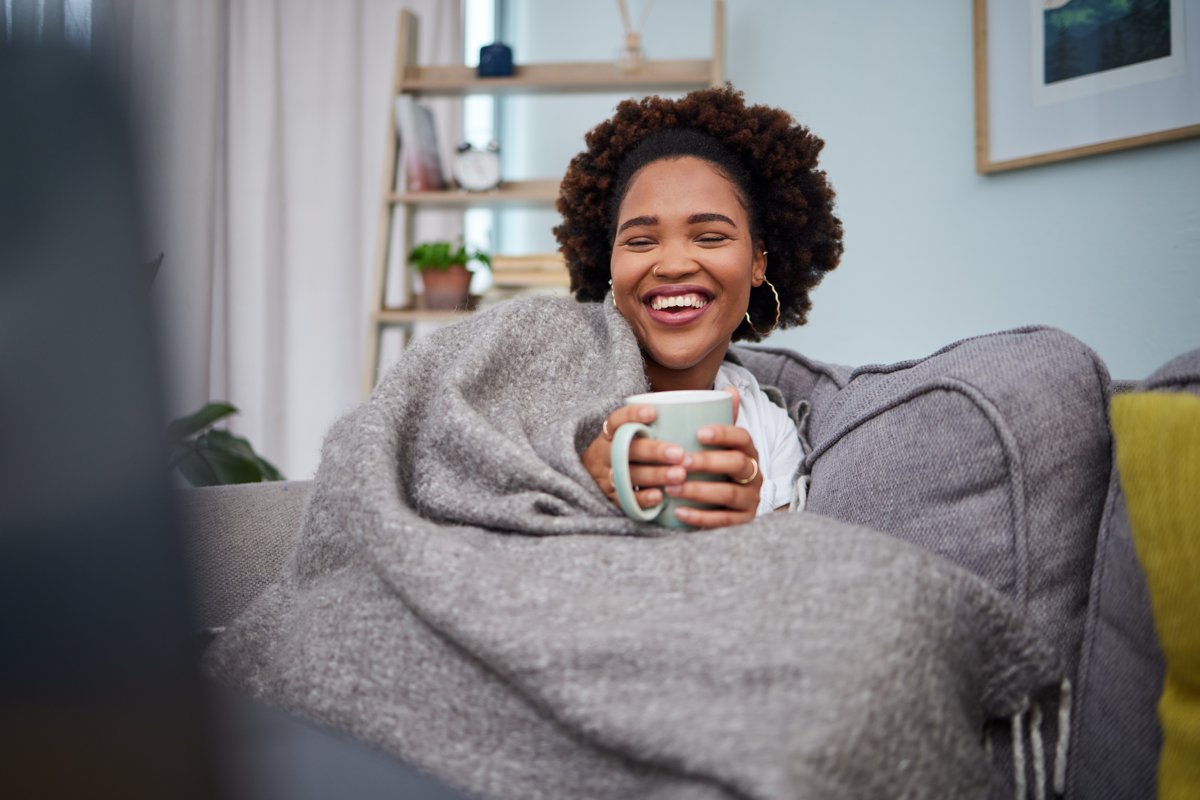 Woman laughing under a blanket with a hot drink on the sofa