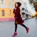Lachende Frau in rotem Mantel und pinken Stiefeletten läuft über eine Straße