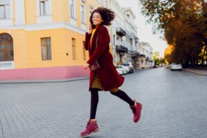 Lachende Frau in rotem Mantel und pinken Stiefeletten läuft über eine Straße