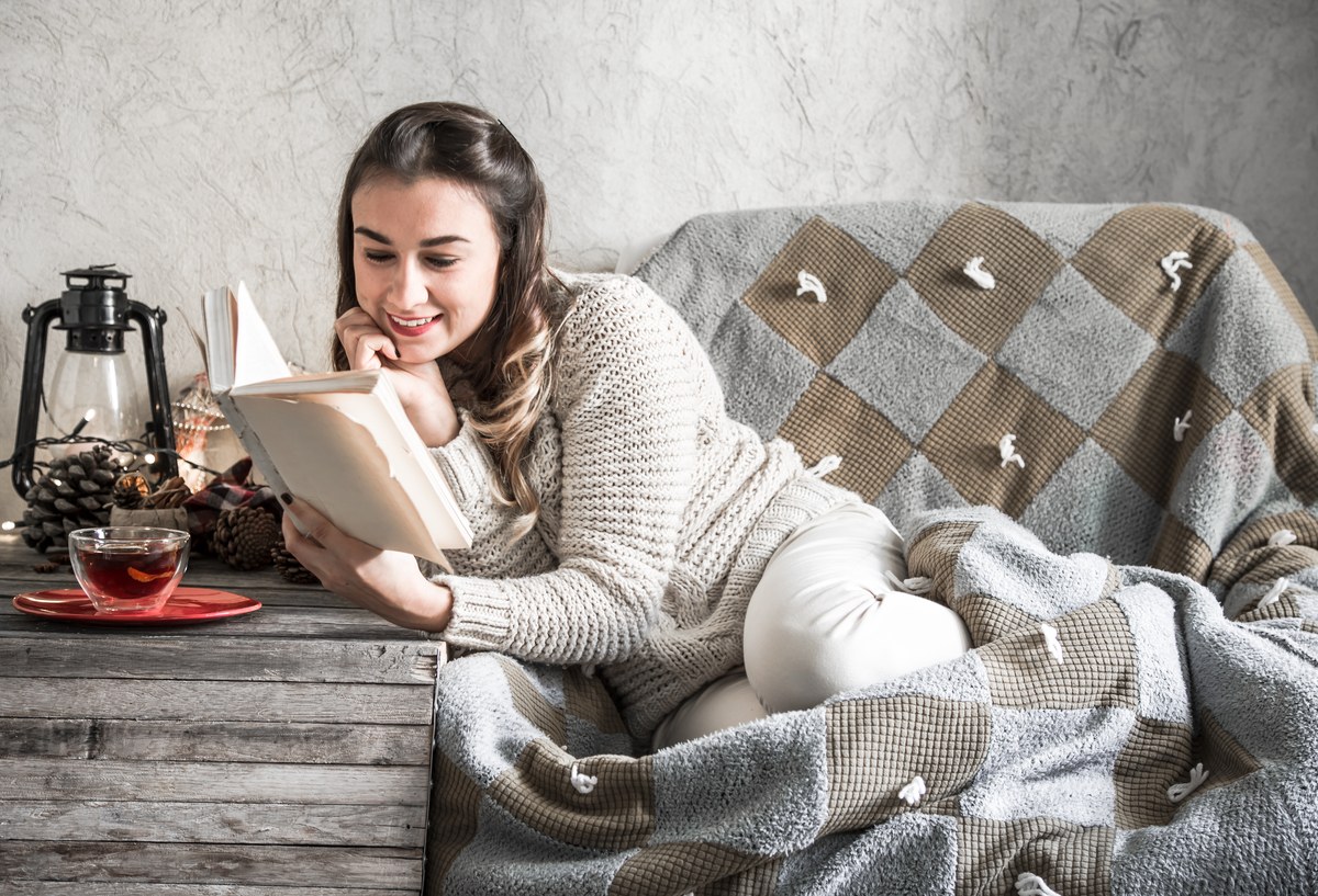 Femme souriante qui lit un livre sur un canapé avec une couverture et une tasse de thé