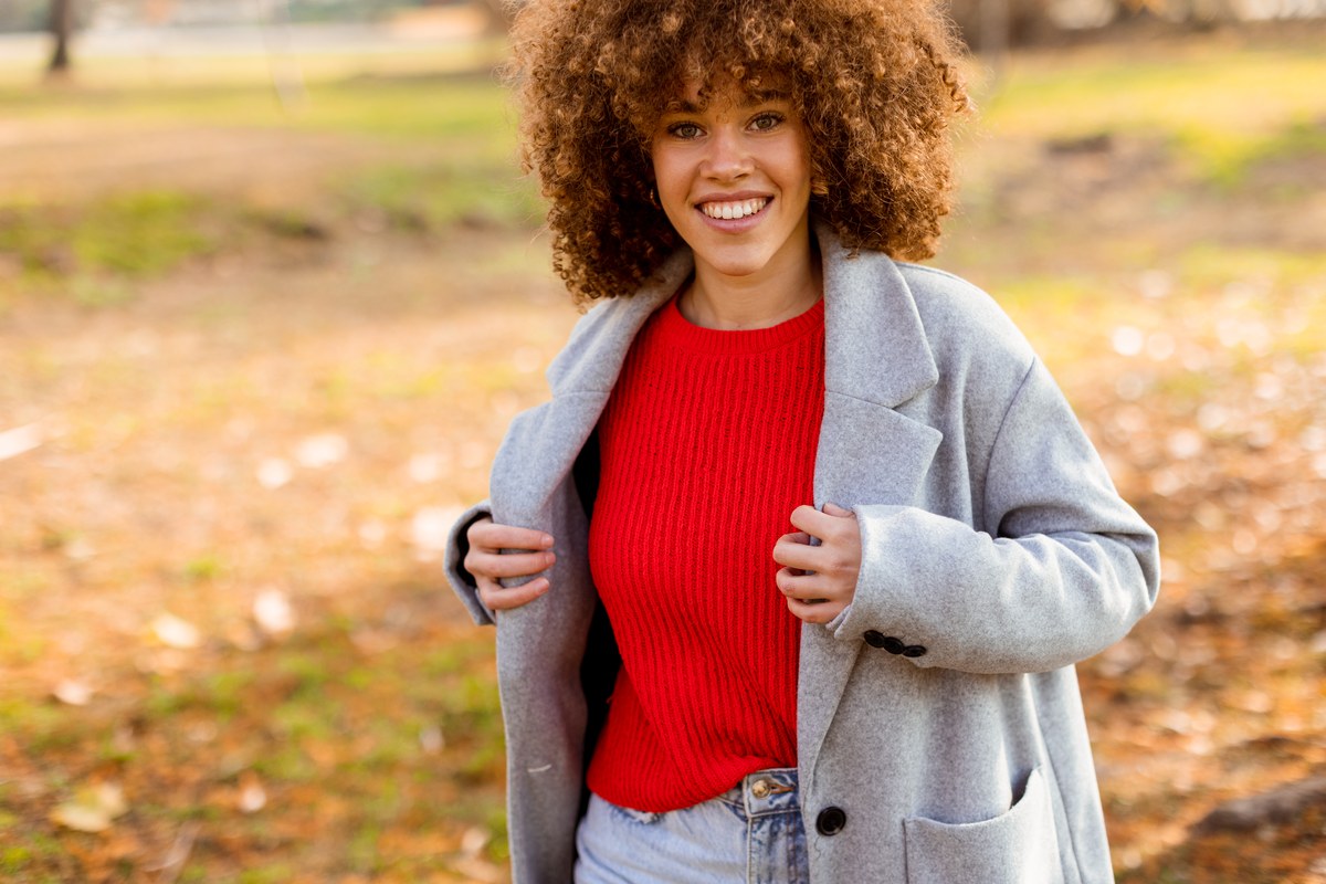 Femme souriante en pull rouge et manteau gris dans un parc en automne