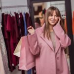 Lachende vrouw in een roze jas met shoppingtassen tussen de kledingrekken