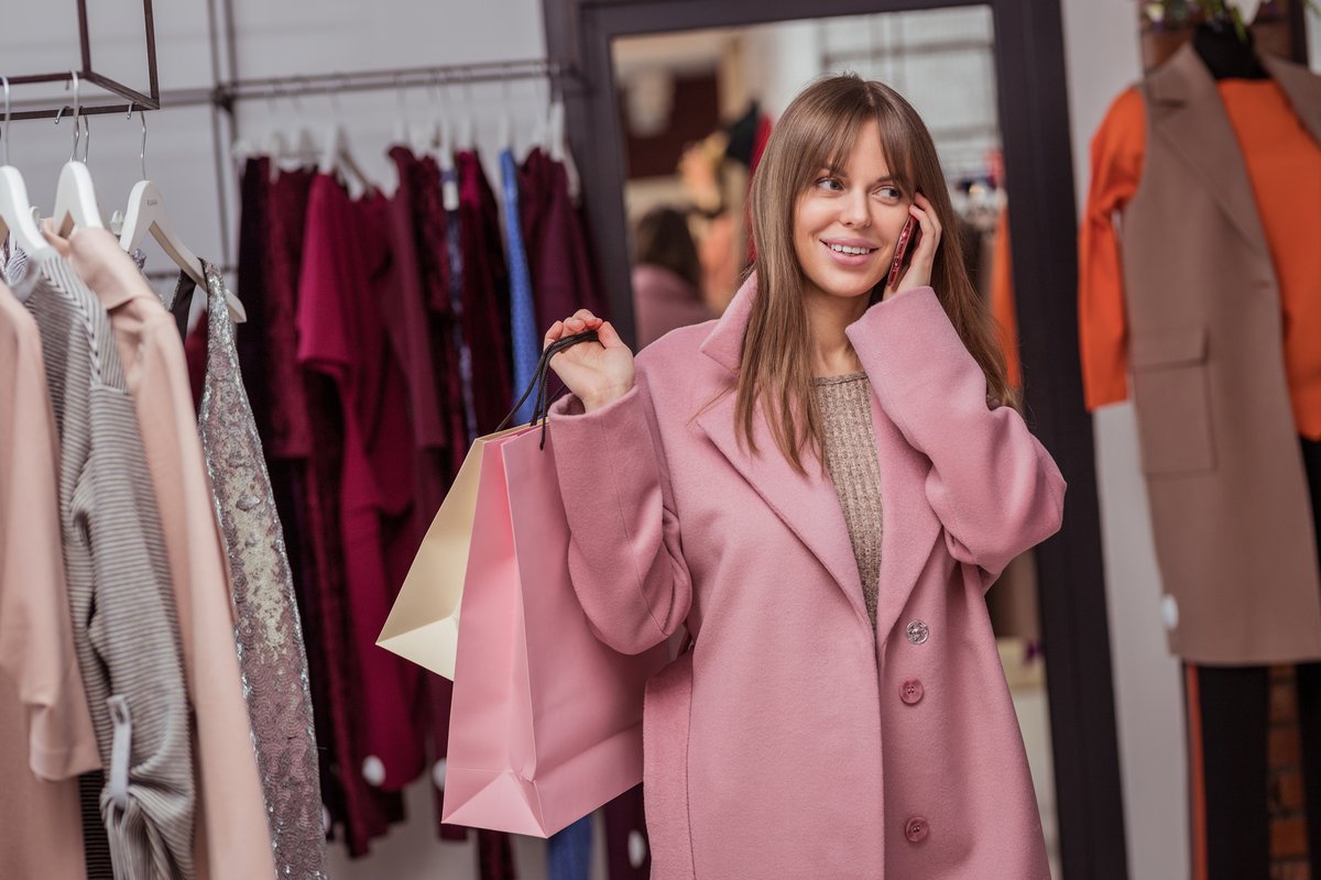 Lachende vrouw in een roze jas met shoppingtassen tussen de kledingrekken