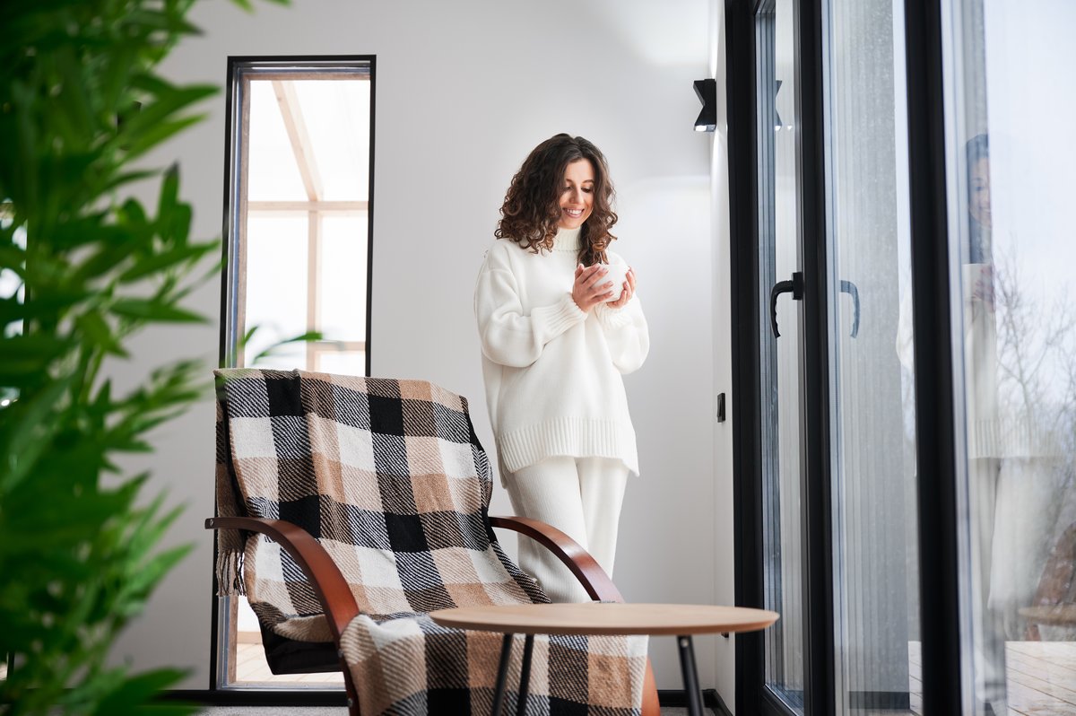 Lachende vrouw in een witte trui drinkt thuis een warme kop thee bij het raam