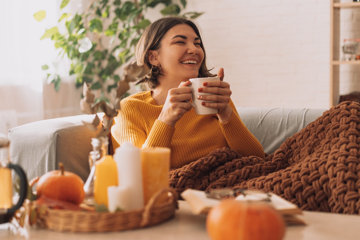 Lachende vrouw met een mok onder een plaid op de bank in de herfst