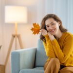 Smiling woman in a yellow jumper holding an autumn leaf on a sofa next to a floor lamp