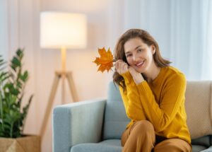 Smiling woman in a yellow jumper holding an autumn leaf on a sofa next to a floor lamp
