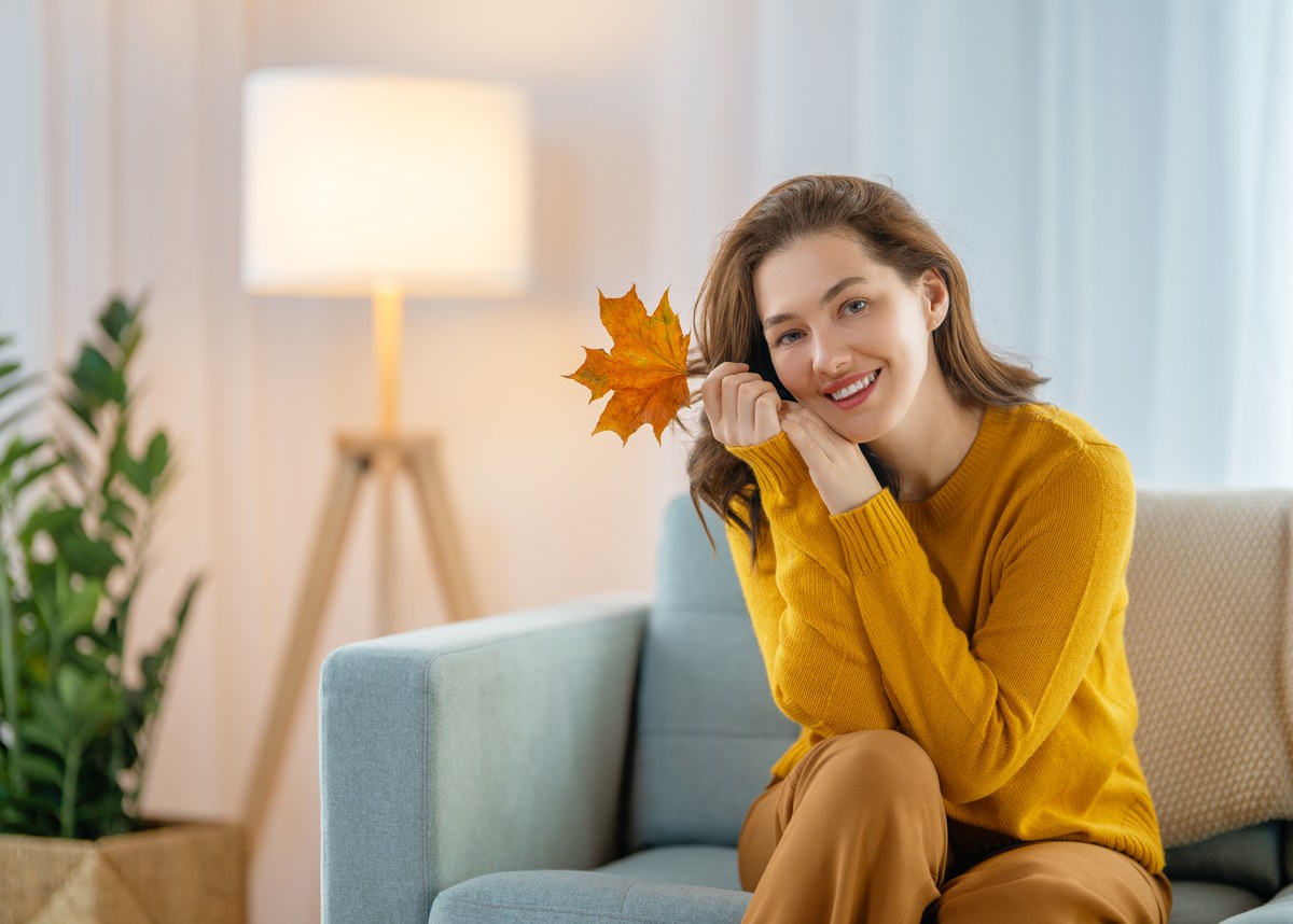 Smiling woman in a yellow jumper holding an autumn leaf on a sofa next to a floor lamp