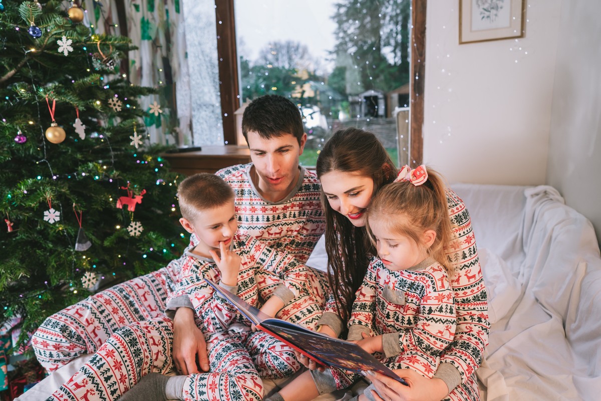 Family in matching Christmas pyjamas reading a book together by the tree