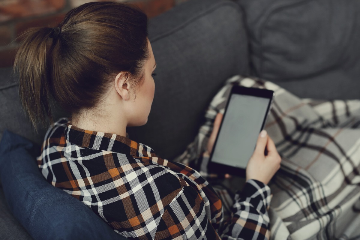 Woman relaxing on a couch under a plaid blanket reading on a tablet