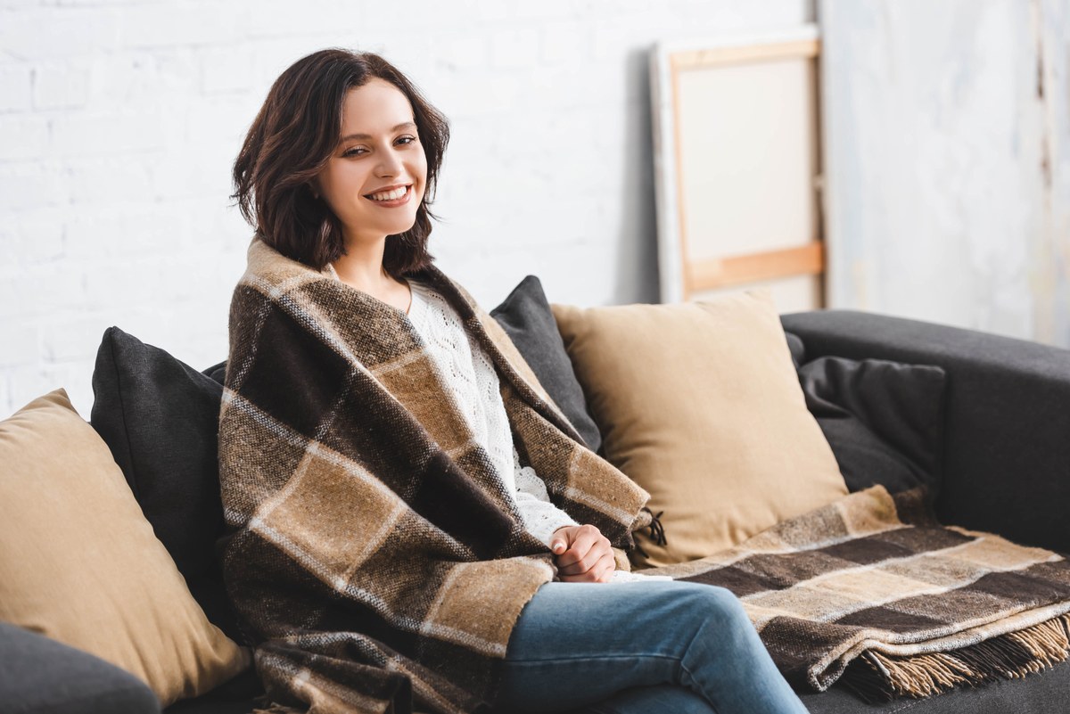 Smiling woman relaxing on a sofa wrapped in a plaid blanket